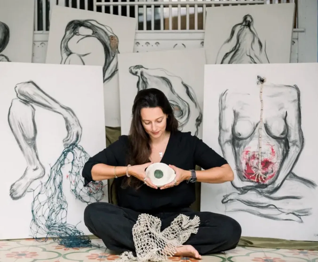 Sofiya Shukhova sitting cross-legged on a patterned tile floor, holding a shallow ceramic bowl with a disc of green sea-worn glass in it. Behind her, large charcoal drawings of human figures lean against the wall; blue fishing net hangs from one, and another carries a red pigment mark low on the torso. Pale netting lies in her lap.